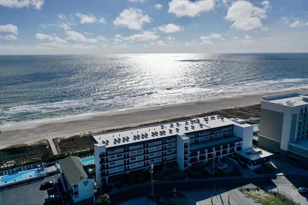 A sweeping exterior view of the ocean, sandy beach, hotel structure, and a community swimming pool. The tranquil setting highlights both the proximity to the beach and the inviting pool area, with gentle waves and soft light conveying a sense of year-round relaxation and fun.