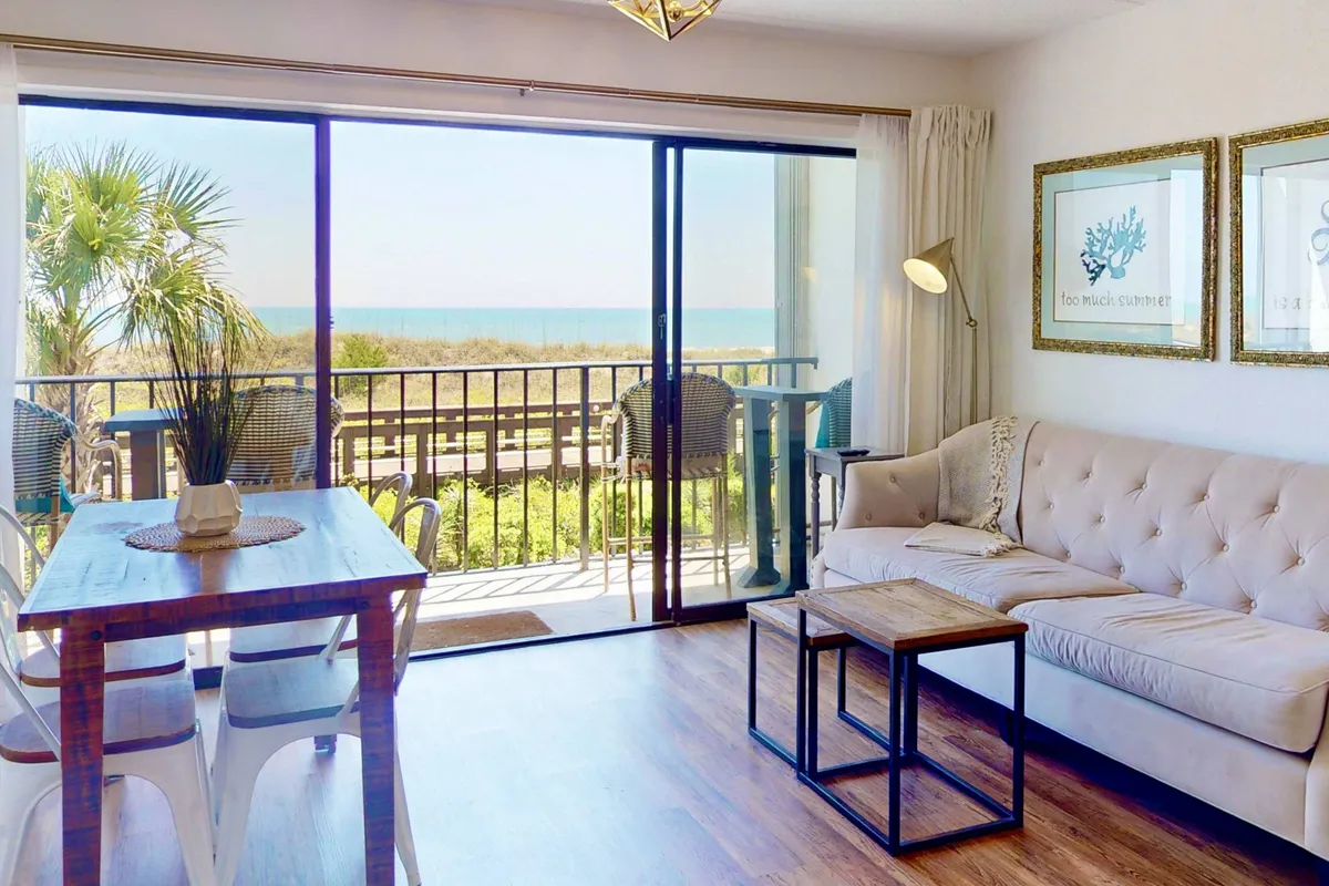 The image showcases a bright, inviting condo interior with large sliding glass doors that open to stunning beach views. A comfortable sofa and a modern dining table are positioned to enjoy the scenery, creating a seamless connection between indoor relaxation and the beautiful Carolina Beach coastline just beyond.