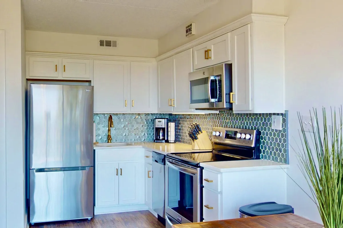 A bright, contemporary kitchen featuring a stainless steel refrigerator, hexagonal tile backsplash, modern stove and microwave, wooden countertops, and a selection of utensils neatly arranged. The layout is open and inviting, creating an ideal space for cooking and meal preparation while staying connected with others nearby.
