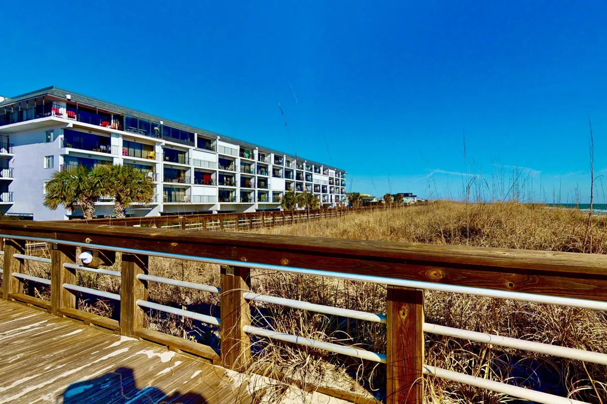 A wooden boardwalk bordered by beach grass leads to a modern beachfront building under a bright blue sky. The pathway invites exploration, with the promise of ocean views and beach access, while the architecture suggests modern amenities and convenience nearby.