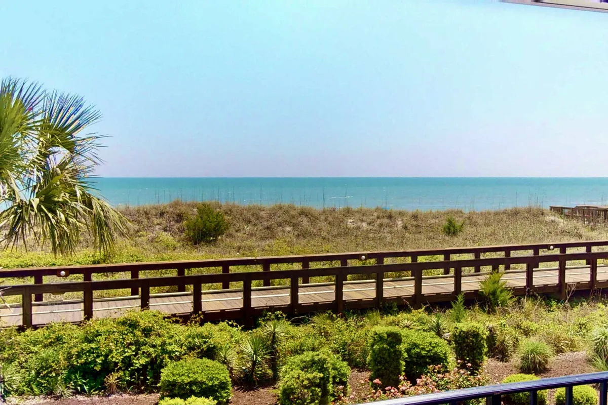 A wide view of the sandy beach meeting the rolling waves of the sea, with the boardwalk visible and lush coastal vegetation framing the scene. People can be seen walking along the shore, sunbathing, and enjoying a tranquil atmosphere.