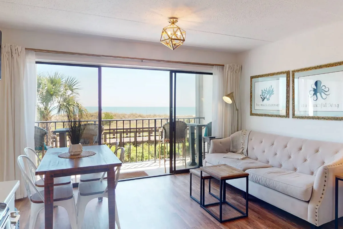 A light-filled dining area with large windows offering an ocean view, a stylish chandelier overhead, a comfortable sofa, and a sizable dining table. This space effortlessly blends natural light, scenic vistas, and a setting ideal for group meals or planning tomorrow's adventures together.