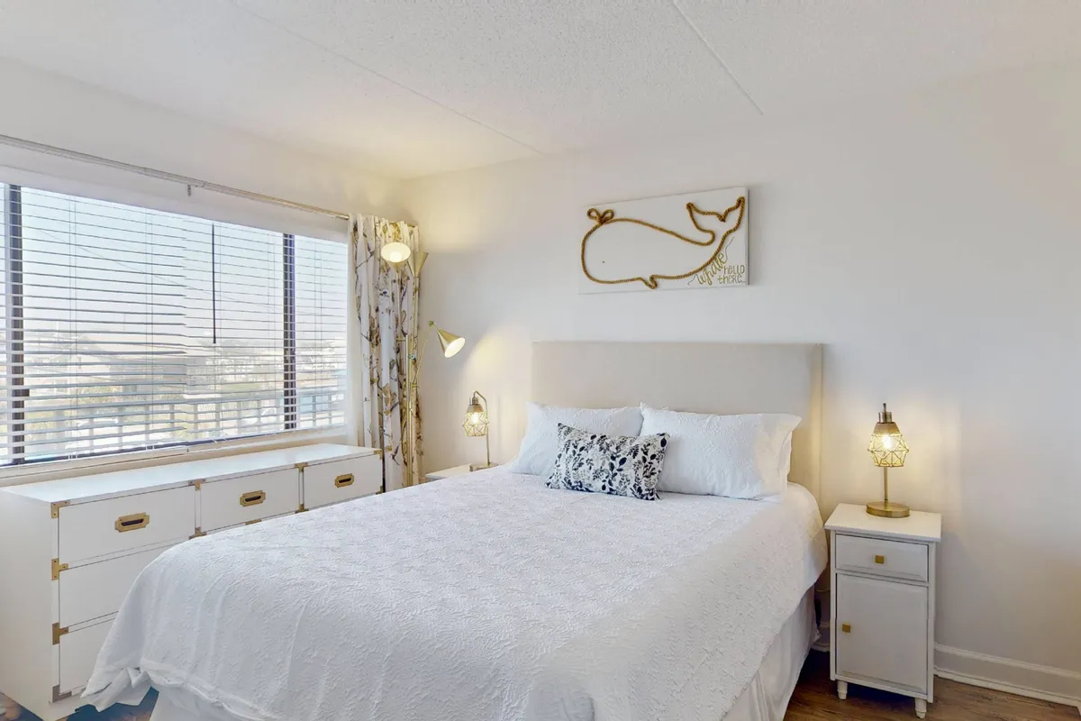 A tranquil bedroom featuring a cozy queen bed dressed in crisp white linens. There is a large window that lets in natural light and offers a glimpse of the outdoors, with soothing whale wall art above the bed and a nightstand with a lit lamp, creating a calm atmosphere.