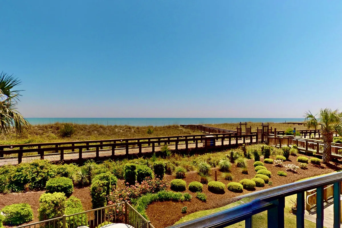 A peaceful view featuring a wooden boardwalk leading through a landscaped garden toward the ocean. The scene is bathed in gentle light, with the serene sea in the background, evoking the allure of sunrise walks and tranquil beach moments.