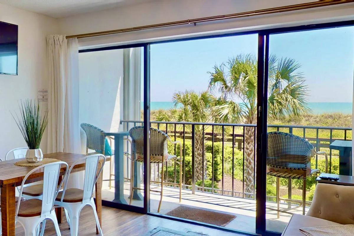 This image captures an open-plan living area with a stylish dining table positioned beside large sliding glass doors. The doors lead out to a private balcony furnished with chairs, all framing a sweeping ocean view with palm trees visible. The room is bathed in natural light, and the tranquil ambiance invites relaxation and togetherness.