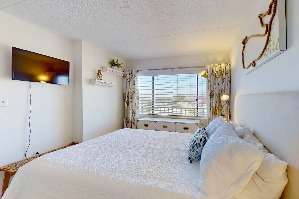 A second inviting bedroom featuring a queen-size bed with plush bedding, a window with curtains allowing gentle daylight, and a TV mounted on the wall. The space is calm and softly lit, ideal for both relaxation and leisure nights in.
