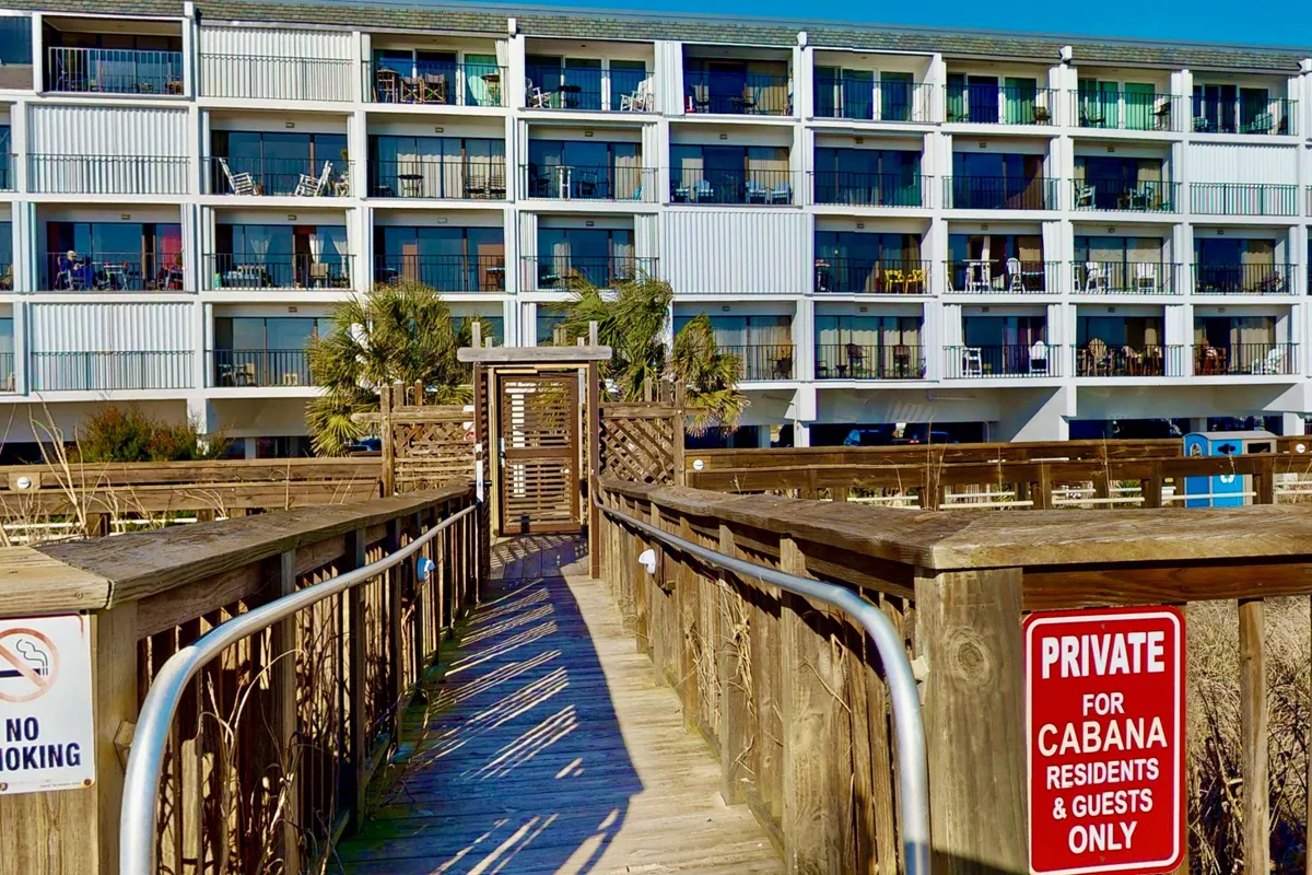 Exterior shot of the property’s building facade, highlighting the wooden walkway (boardwalk), palm trees, and clear signage. Residents and guests are seen enjoying the walk or relaxing on balconies, emphasizing the relaxed and accessible vibe.