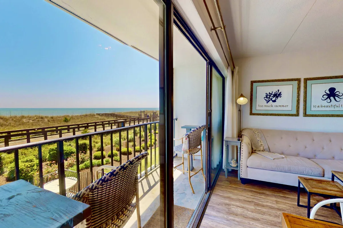 A cozy, inviting seating area arranged to face a balcony with an expansive ocean view. The decor is serene and modern, with wall art accenting the space, and comfortable furniture inviting relaxation as natural light floods the room.