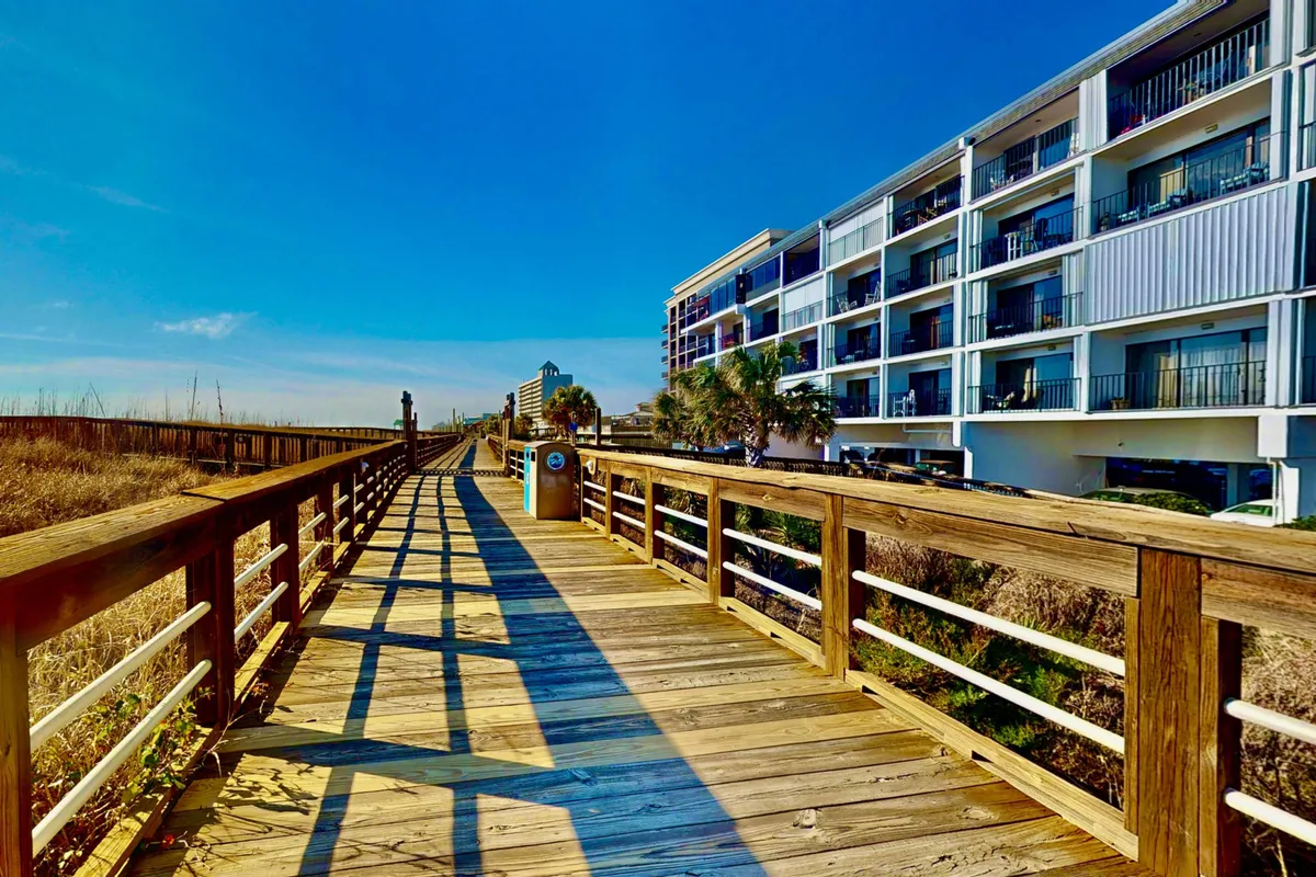 This image presents a stunning exterior perspective with a wooden boardwalk leading directly to the ocean. Palm trees frame the path, and an apartment building is visible, emphasizing both the proximity to the beach and the inviting, walkable atmosphere. The view captures the vibe of early morning sunrises and the community's friendly energy.