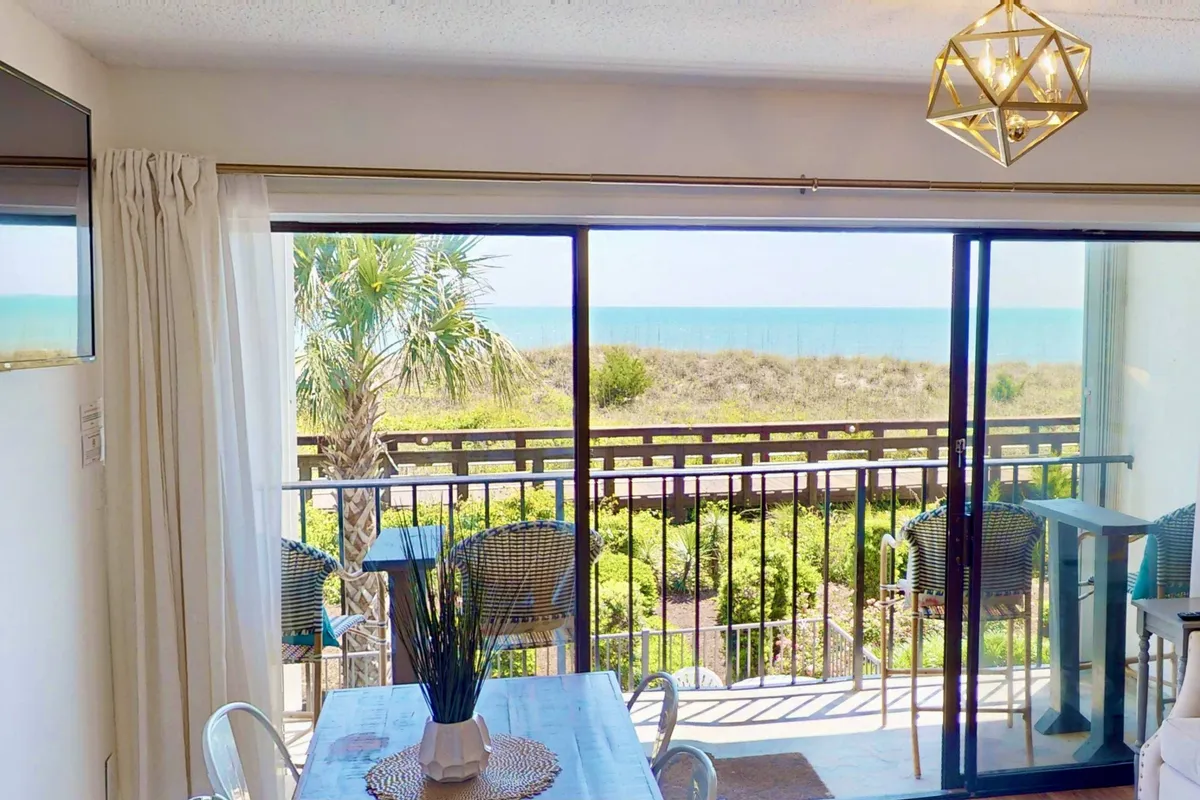 A spacious interior with a large glass door opening onto a balcony; inside, a dining table with a decorative centerpiece is set for meals with an ocean backdrop. The cozy balcony seating area invites guests to relax while soaking in the view and enjoying the breeze.