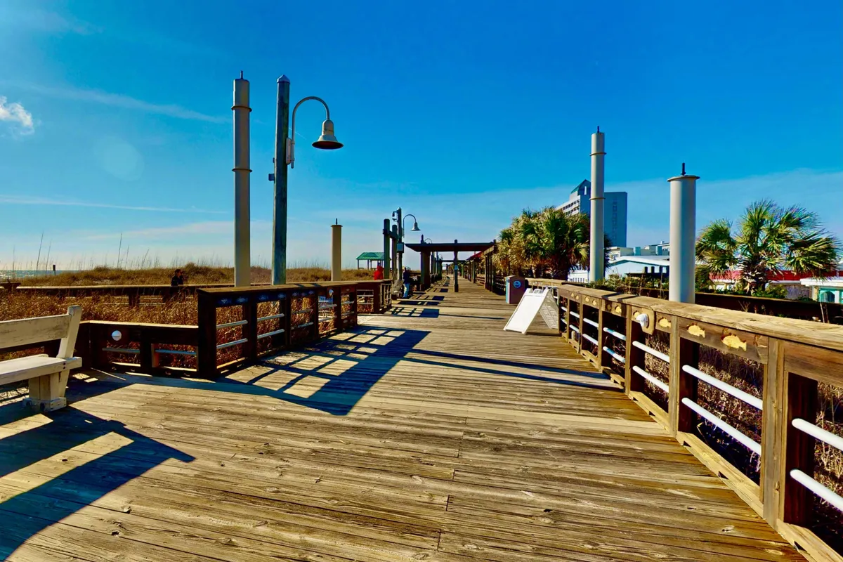 A lively boardwalk with wooden planks, street lamps, benches, and beach vegetation leading towards the ocean. Buildings and festival elements are visible in the background, suggesting community activities and events.