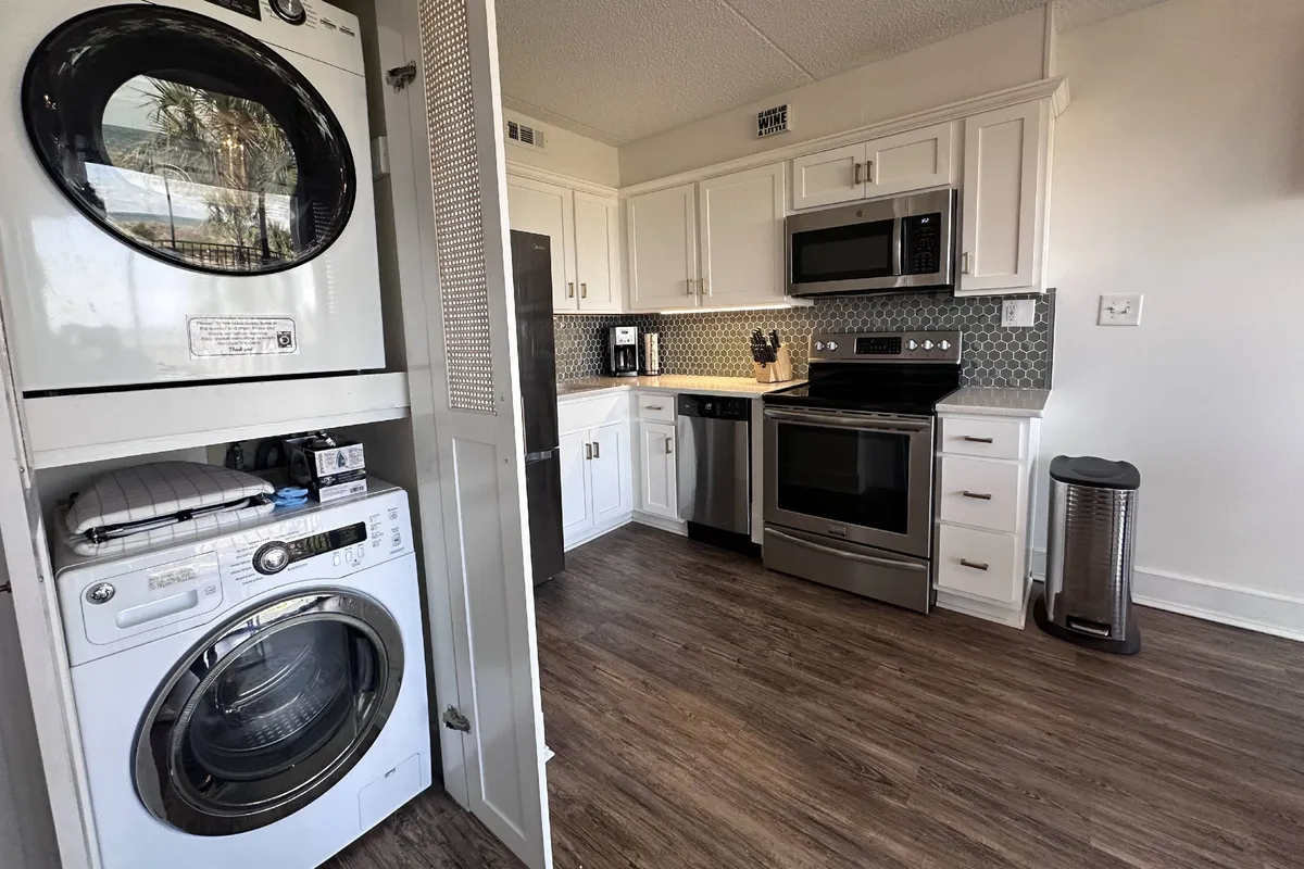 An interior view of the condo’s laundry area with a modern washer and dryer tucked into a sleek kitchen, featuring stainless steel appliances and cabinetry. The space looks tidy and efficient, promising convenience for guests or residents.