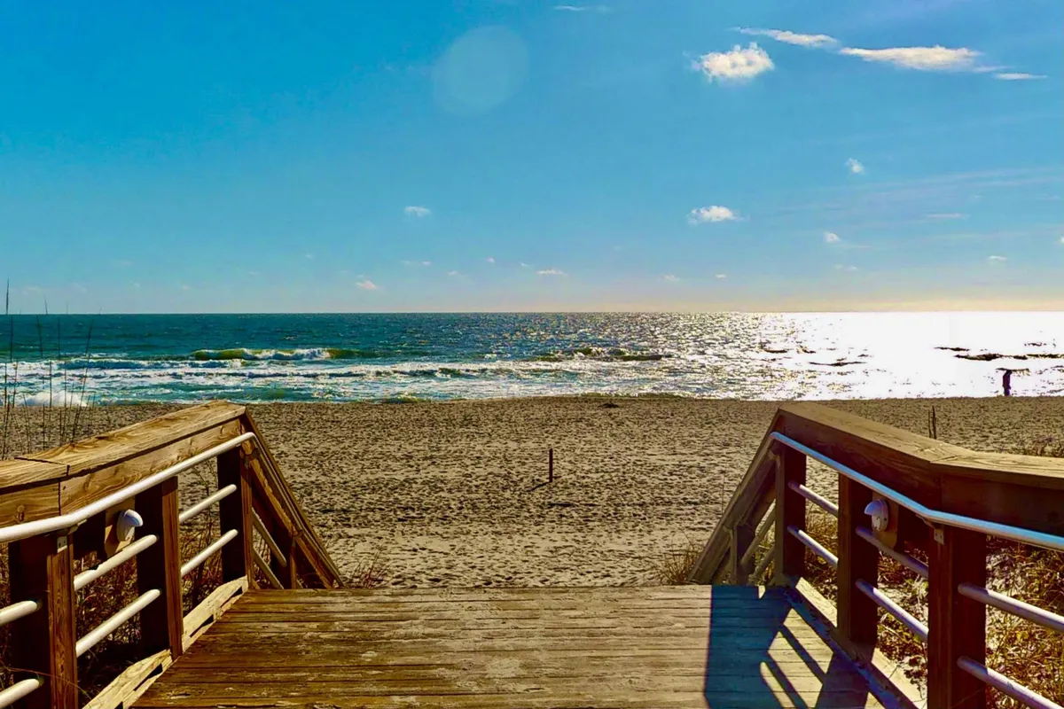 A sweeping view of a wooden walkway extending toward a wide beach and open ocean, under a clear sky. The scene is tranquil, hinting at various coastal activities like walking, sunbathing, surfing, and fishing. The expansive shoreline and inviting waves evoke a sense of adventure and exploration.